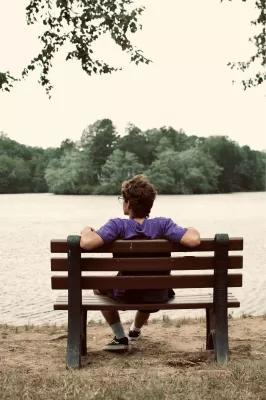Un home d'esquenes seu a un banc a la vora d'un llac.